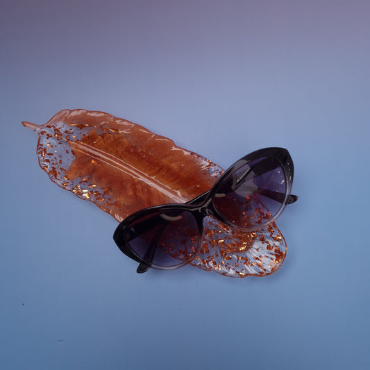 Bandeja em resina epóxi formato pena, tom cobre com partículas douradas, vista frontal com óculos pousados.