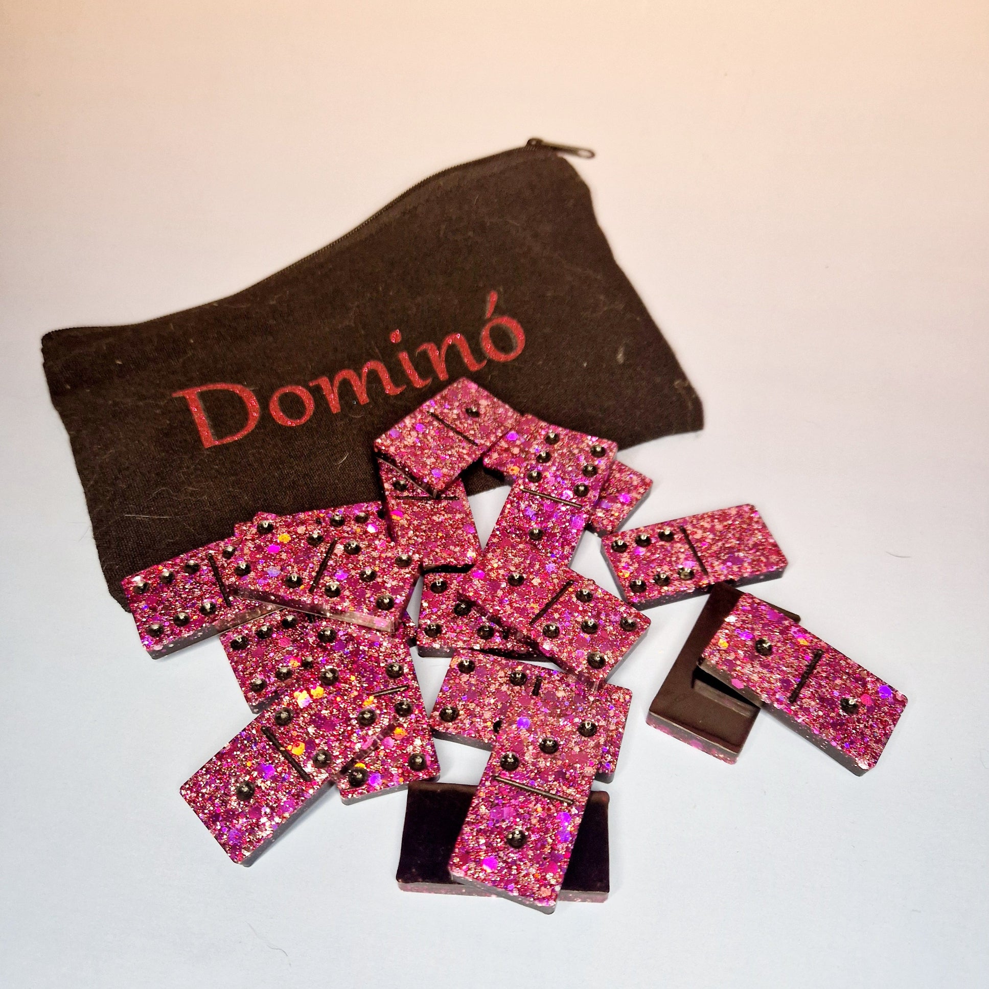 Dominó artesanal em resina rosa com glitter, conjunto completo disposto à frente de um estojo em tecido escuro com a palavra “Dominó”.