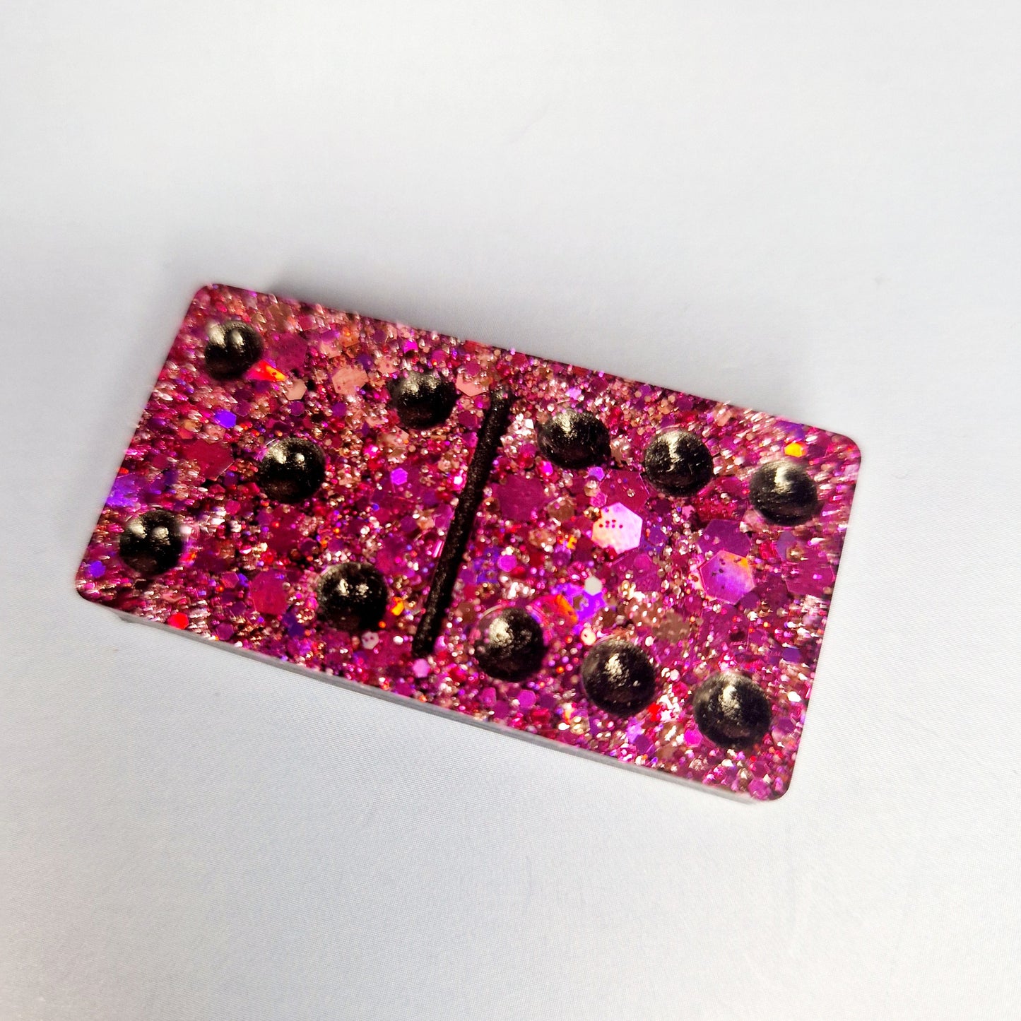 Peça individual de dominó artesanal em resina rosa glitter com pontos em relevo pretos, vista aproximada sobre fundo claro.
