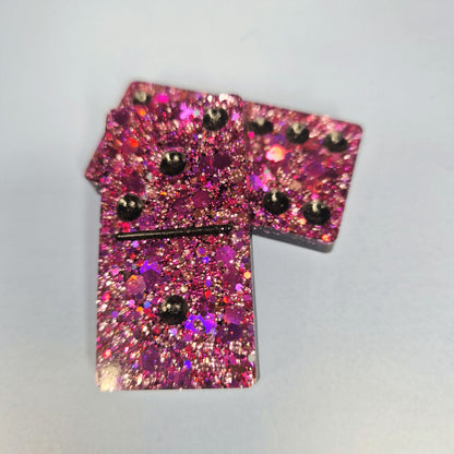 Duas peças de dominó artesanal em resina rosa com glitter intenso, sobrepostas, com detalhe dos pontos em relevo.