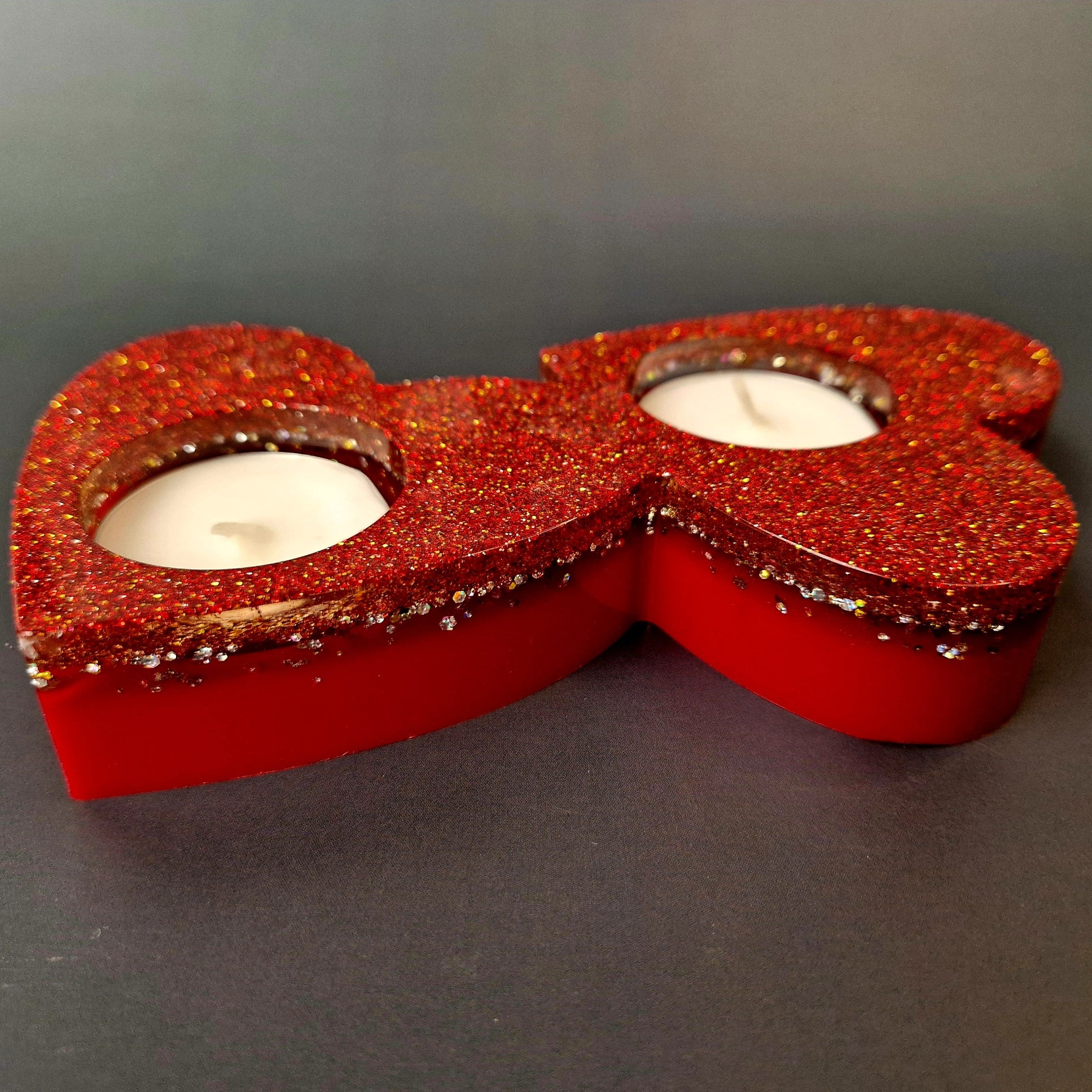 Porta-velas duplo em forma de coração em resina epóxi vermelha com glitter dourado, visto de cima, com duas velas brancas.