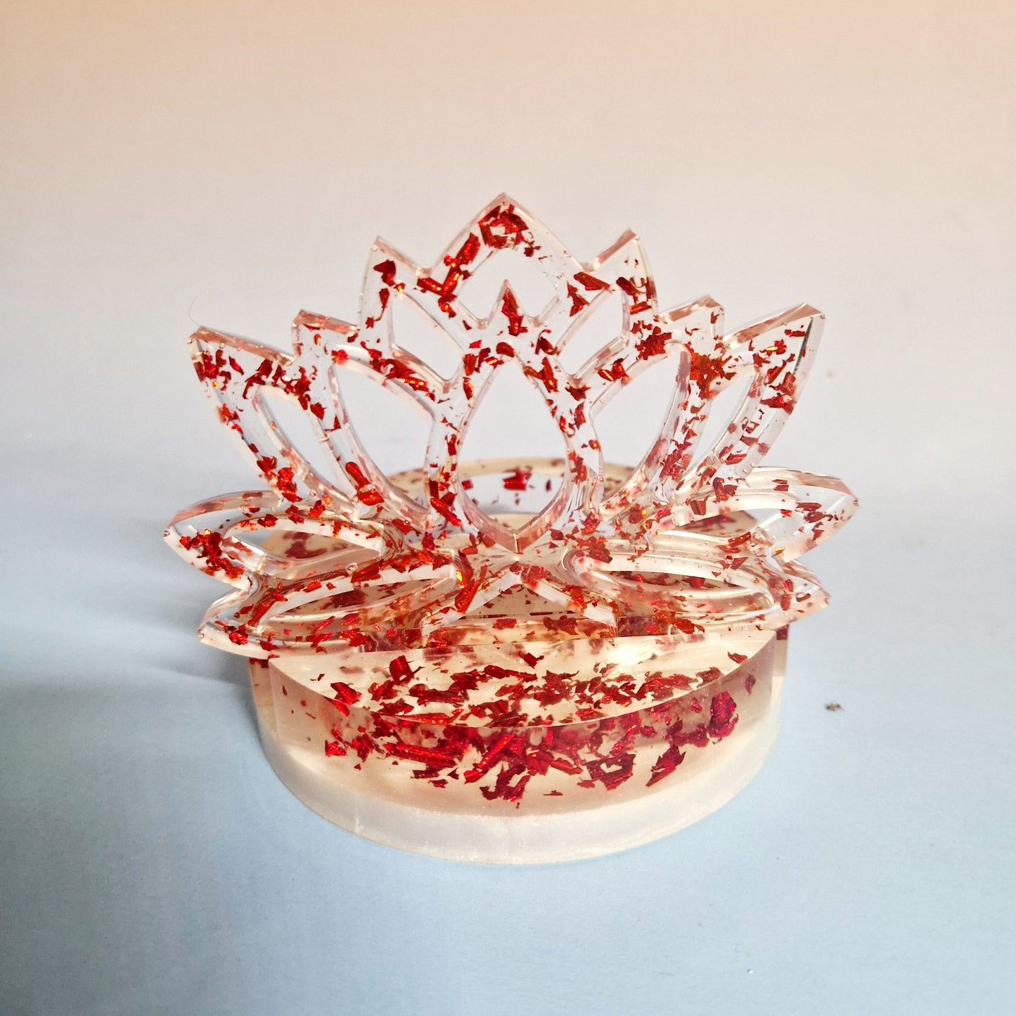 Suporte de vela em resina transparente com silhueta de Flor de Lótus e flocos metálicos vermelhos em fundo neutro.