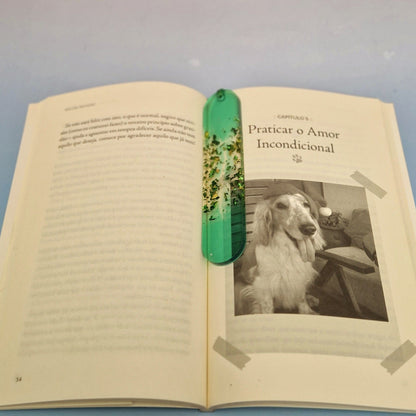 Marcador de página artesanal em resina verde translúcida com glitter dourado, colocado em livro aberto para marcar a leitura.