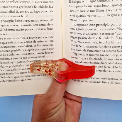 Anel de leitura em resina vermelha translúcida com glitter dourado, encaixado no centro do livro para facilitar a leitura com uma mão.