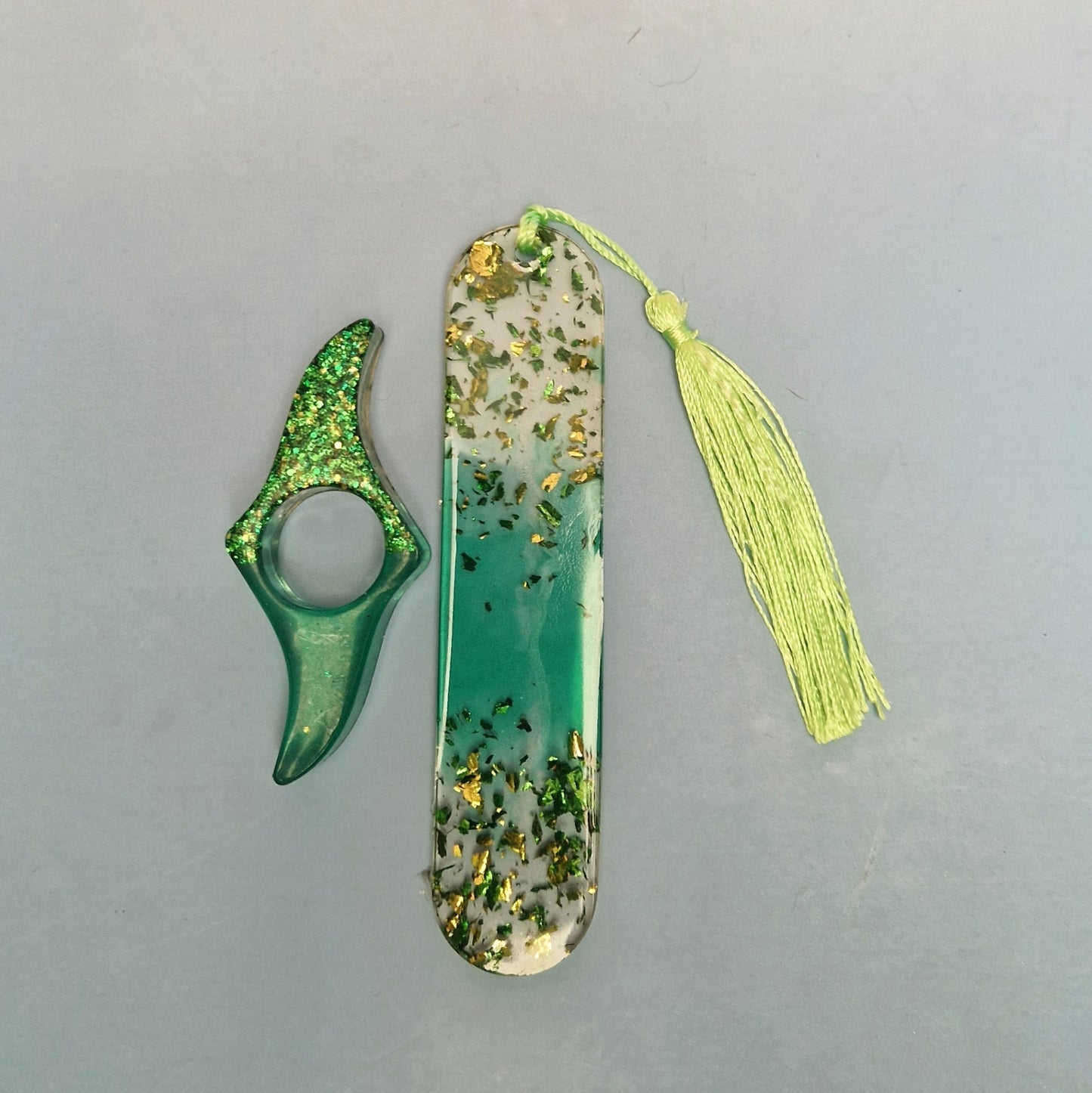 Marcador de página em resina verde translúcida com efeito degradê, folhas douradas decorativas e tassel verde claro, ao lado de acessório artesanal combinando.