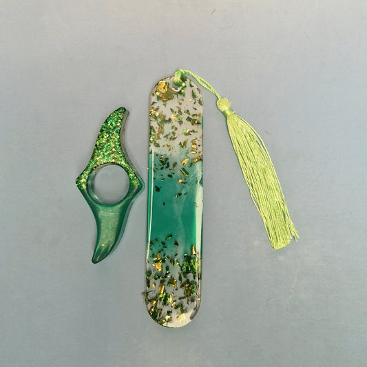 Marcador de página em resina verde translúcida com efeito degradê, folhas douradas decorativas e tassel verde claro, ao lado de acessório artesanal combinando.