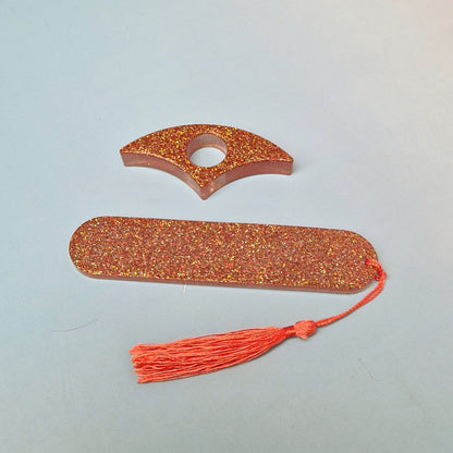 Conjunto artesanal em resina com glitter dourado e vermelho, marcador de página retangular e acessório curvo com furo central, acompanhado de tassel Salmao
