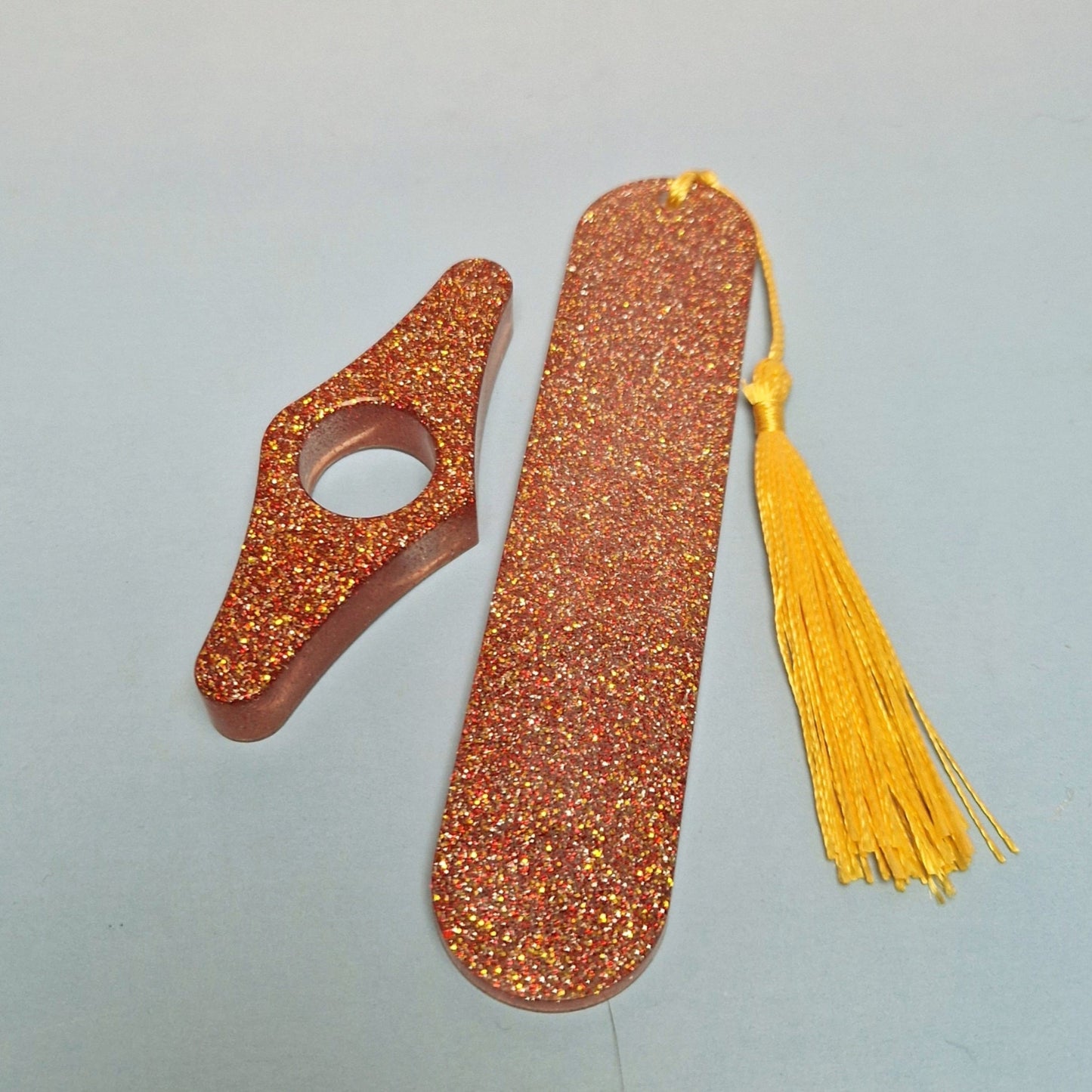 Marcador de página artesanal em resina com glitter dourado e vermelho, formato alongado, tassel amarelo e acessório decorativo curvo combinando.